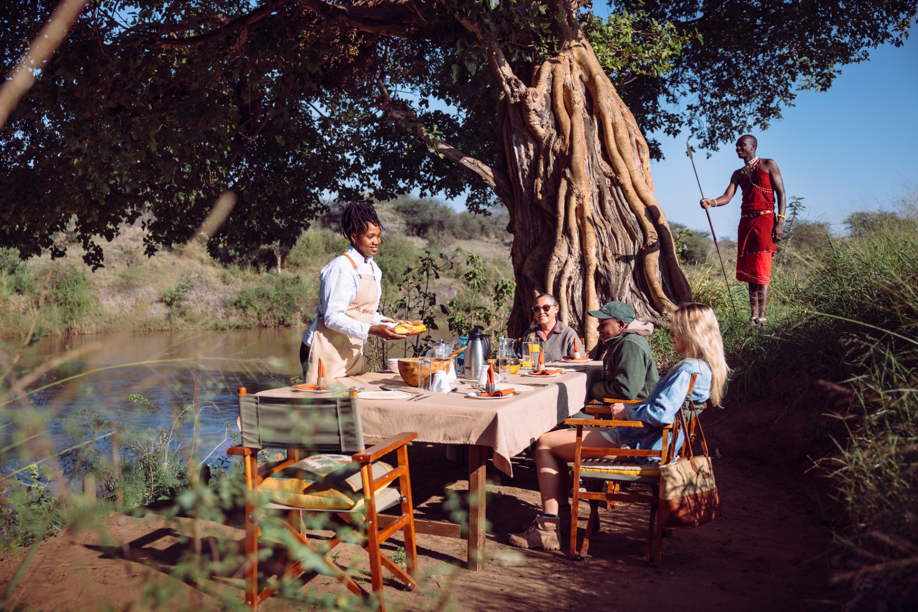Chef serving breakfast to guests beside a river, with a Maasai guide standing nearby